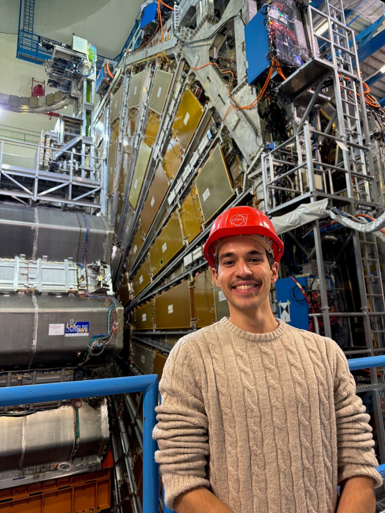Sina wearing a CERN hard hat in front of particle detector equipment at CERN.